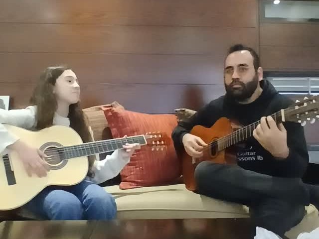 Un élève et Elias jouent de la guitare classique ensemble sur un canapé, à la maison