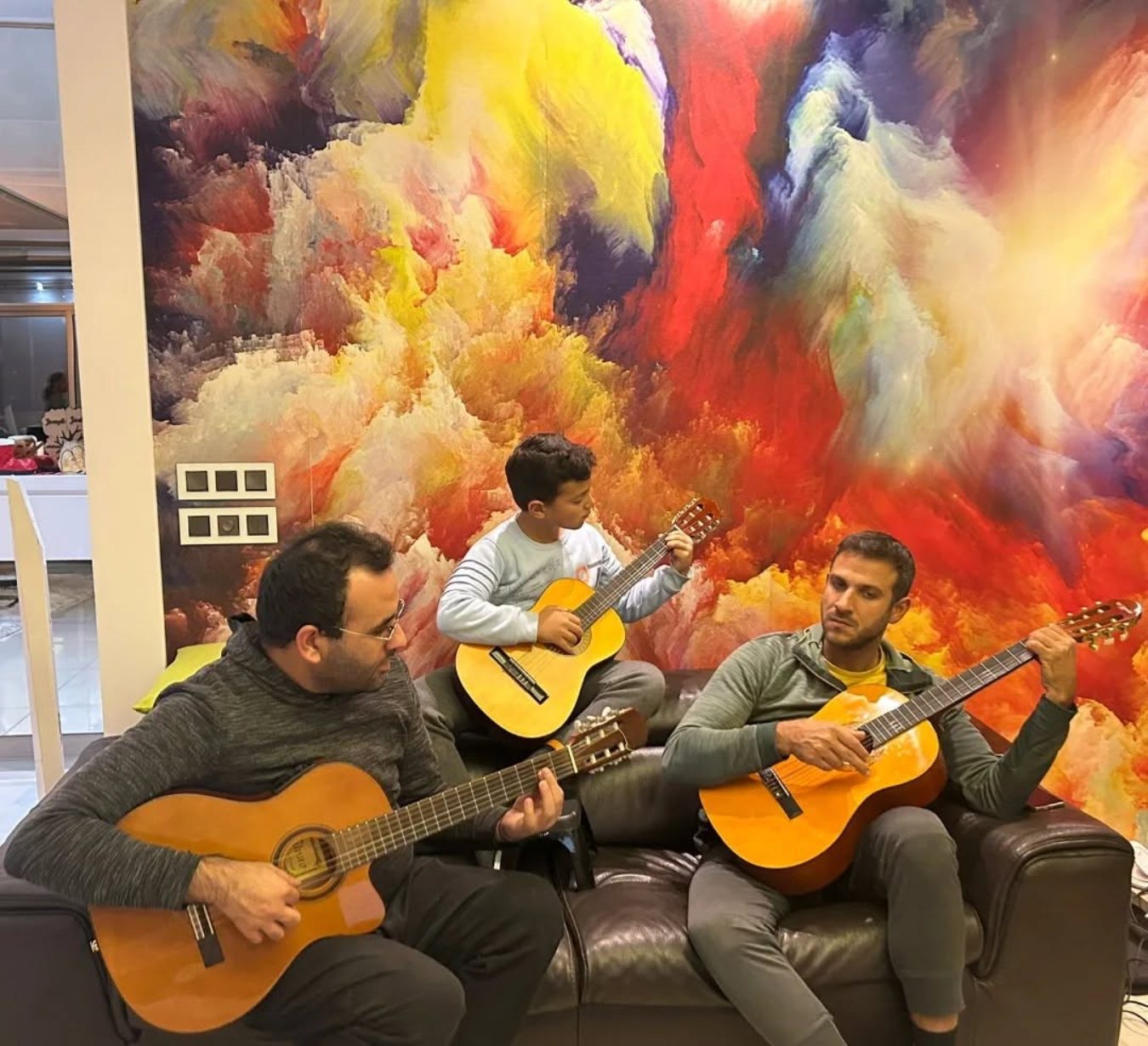 Two teachers and a young student playing classical guitars together on a sofa in front of a colourful painted wall